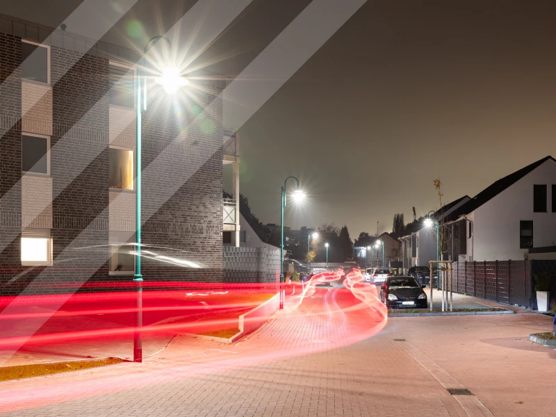 Nächtliche Straßenszene mit moderner LED-Straßenbeleuchtung: Leuchten erhellen eine Wohnstraße, während rote Lichtspuren vorbeifahrender Fahrzeuge Bewegung und Dynamik zeigen.