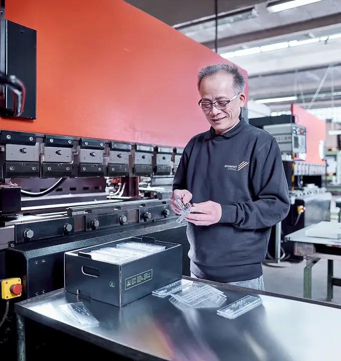 ein Schmidt-Strahl Mitarbeiter in der Produktion prüft Bauteile vor einer Maschine, während vor ihm sortierte Komponenten auf einem Arbeitstisch liegen