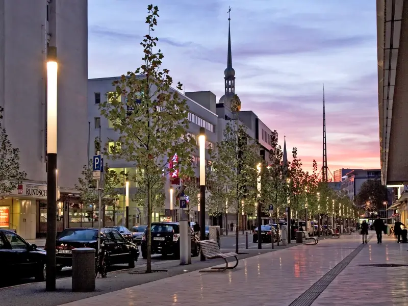 Belebte Innenstadtstraße in der Dämmerung mit modernen, warm leuchtenden Straßenleuchten, parkenden Autos, jungen Bäumen und einer Kirche mit Turm im Hintergrund unter farbigem Abendhimmel
