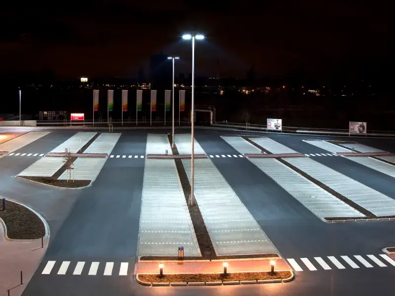 Großer, nahezu leerer Parkplatz bei Nacht mit klar strukturierten Stellflächen, hell ausgeleuchtet durch hohe LED-Mastleuchten, mit Fahrbahnmarkierungen und vereinzelter Umgebungsbeleuchtung im Hintergrund