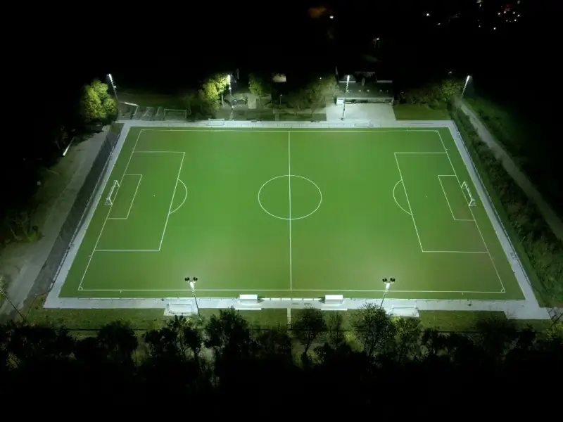 Vollständig ausgeleuchteter Fußballplatz des BV Wevelinghoven bei Nacht aus der Vogelperspektive, mit gleichmäßig hellem Spielfeld, Flutlichtmasten und umliegenden Wegen sowie Grünflächen