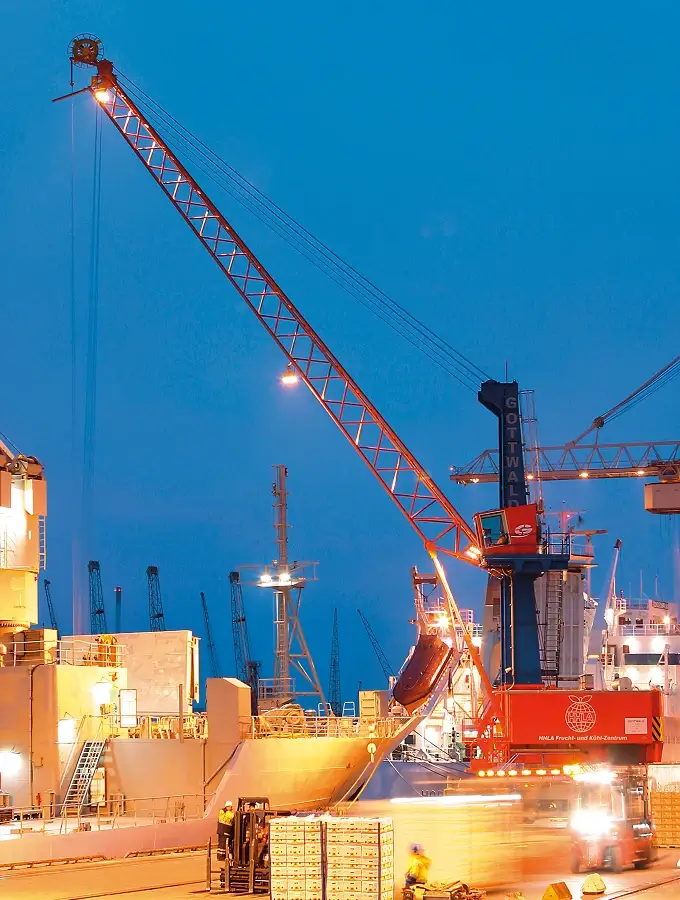 Beleuchteter Hafenkran vom Typ Gottwald beim nächtlichen Verladen von Fracht im Hamburger Hafen, mit Schiffen, Containern und Fahrzeugen im Hintergrund