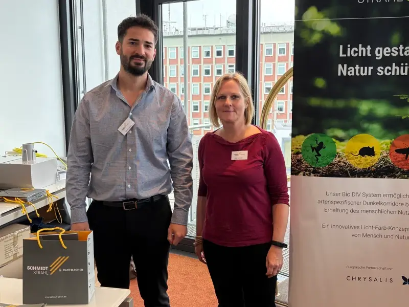 Silke Kesiciler und Serhat Kurnaz präsentieren Schmidt-Strahl auf der Mitgliederversammlung der Überwachungsgemeinschaft Gleisbau (ÜGG) in Hamburg. Neben einem Roll-up zum Thema Naturschutz und Lichtplanung stehen Informationsmaterialien und Produkte für die Bahninfrastruktur bereit. Im Hintergrund sind die Fensterfront des Veranstaltungsortes und Gebäude des Hamburger Hafenumfelds zu sehen.